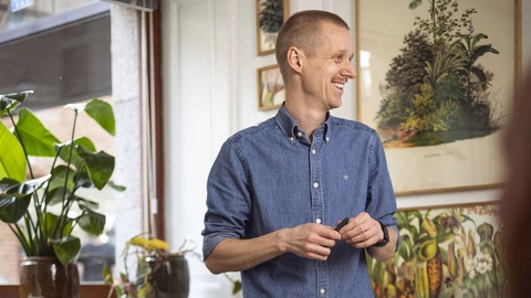 En smilende mand i en blå skjorte præsenterer foran en gruppe mennesker i et lyst rum med planter og farverig vægkunst.